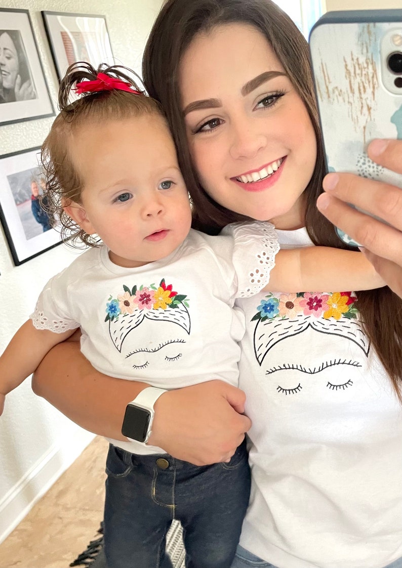 Pu&ograve; includere: Una donna e una bambina piccola indossano magliette bianche abbinate con un'immagine ricamata di Frida Kahlo. La bambina indossa jeans blu e la donna tiene la bambina. La donna indossa una maglietta bianca con bordi in pizzo.