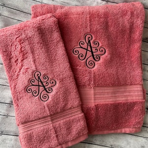 May include: Two pink bath towels with black embroidered cursive letter 'A's. The towels are folded and stacked on top of each other.