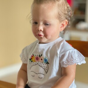 Pu&ograve; includere: Una bambina che indossa una maglietta bianca con un'immagine ricamata di Frida Kahlo. La maglietta ha un bordo di pizzo bianco sulle maniche. La bambina indossa jeans blu e ha un fiocco rosso nei capelli.