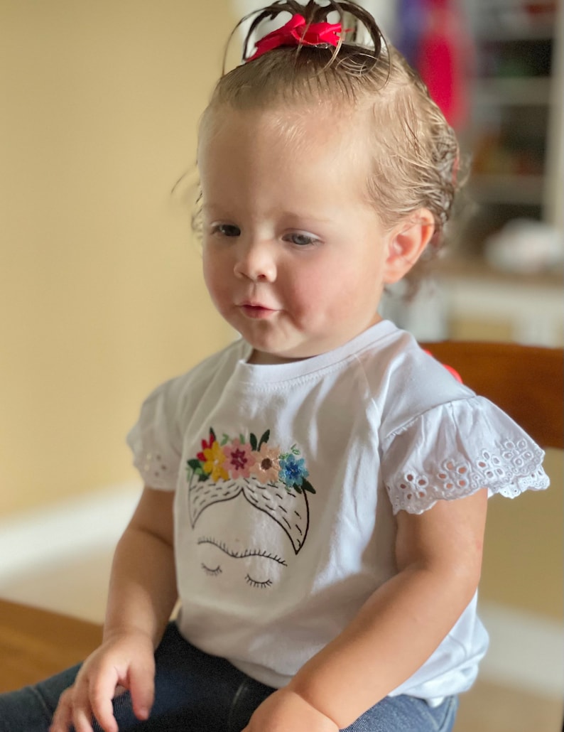 Pu&ograve; includere: Una bambina che indossa una maglietta bianca con un'immagine ricamata di Frida Kahlo. La maglietta ha un bordo di pizzo bianco sulle maniche. La bambina indossa jeans blu e ha un fiocco rosso nei capelli.