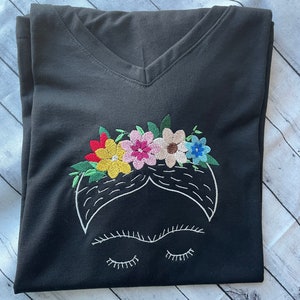 Pu&ograve; includere: Maglietta nera a collo a V con un'immagine ricamata di Frida Kahlo con una corona di fiori. I fiori sono rossi, gialli, rosa, verdi e blu.