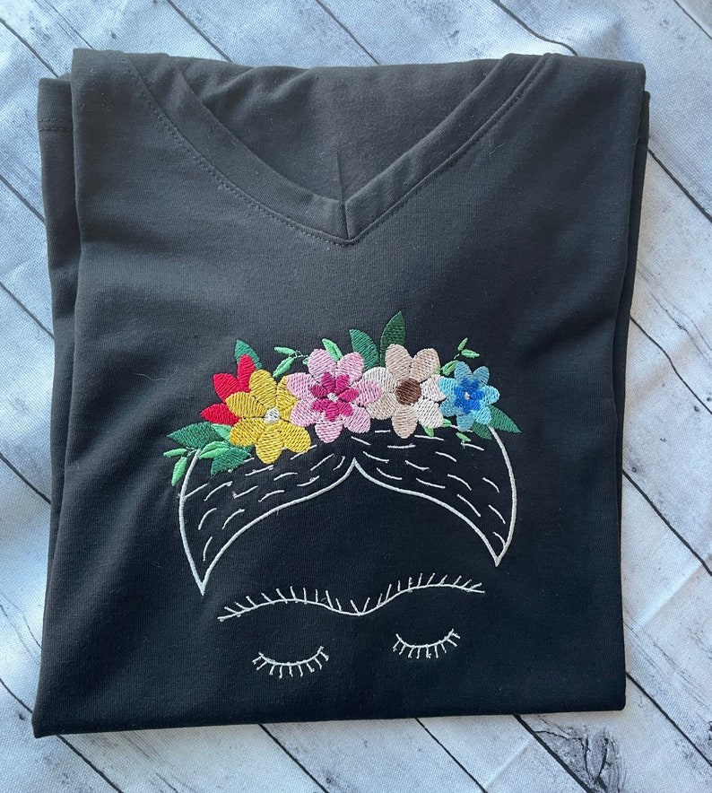 Pu&ograve; includere: Maglietta nera a collo a V con un'immagine ricamata di Frida Kahlo con una corona di fiori. I fiori sono rossi, gialli, rosa, verdi e blu.