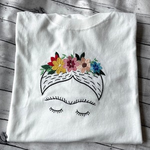 Pu&ograve; includere: Maglietta bianca con un'immagine ricamata di Frida Kahlo con una corona di fiori. La corona &egrave; fatta di fiori colorati in tonalit&agrave; di rosso, giallo, rosa e blu.