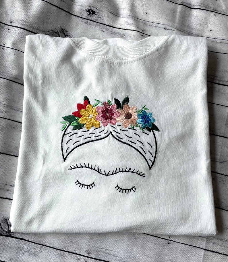 Pu&ograve; includere: Maglietta bianca con un'immagine ricamata di Frida Kahlo con una corona di fiori. La corona &egrave; fatta di fiori colorati in tonalit&agrave; di rosso, giallo, rosa e blu.