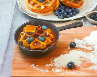 May include: A cast iron skillet with a waffle candle, a stick of butter, and blue berry candles. The skillet is on a wooden cutting board with powdered sugar scattered around it.
