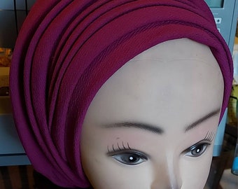 May include: A deep magenta turban-style head covering, artfully draped and wrapped. The fabric has a textured appearance, and the head covering is displayed on a mannequin head. The background includes a sewing machine.