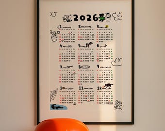 May include: A framed 2026 calendar with a black border. The calendar features hand-drawn illustrations and the months of the year. Each month has a unique design, with dates in red and black. The calendar is hanging on a wall.