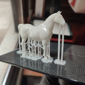 May include: A white 3D printed horse model with support structures still attached. The horse is standing on a black and white patterned surface.