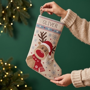 Puede incluir: Un calcetín navideño de tela beige con un diseño de punto de cruz de un reno con gorro y bufanda rojos. El calcetín incluye bordes decorativos azules y rojos, pequeños corazones rojos, patrones de copos de nieve y el nombre OLIVER bordado.