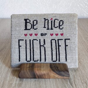 Könnte beinhalten: Ein beigefarbener Canvas mit schwarzer Stickerei, auf dem "Be nice or F*** off" mit roten Herzen zwischen den Wörtern steht. Der Canvas steht auf einem Holzständer.
