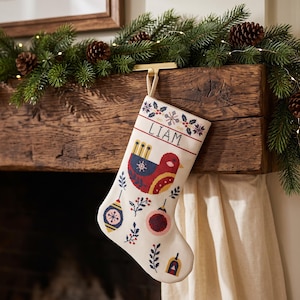 Pode incluir: Uma meia de Natal cor de creme com um design em ponto cruz de um pássaro vermelho, ornamentos decorativos e ramos de azevinho. O nome LIAM está bordado com fio preto perto da borda superior, que inclui uma alça para pendurar a peça.
