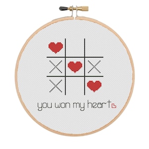 Peut inclure: Un cerceau de broderie blanc avec un motif de point de croix noir et rouge. Le motif représente un jeu de tic-tac-toe avec des cœurs à la place des X et des O. Le texte "you won my heart" est brodé sous le tableau.
