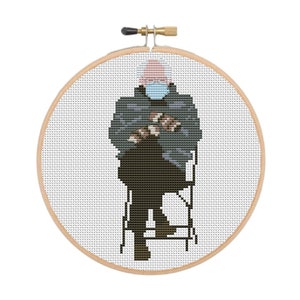 May include: Cross-stitch embroidery of a person wearing a blue face mask and sitting on a chair. The person is wearing a dark jacket and has white hair.