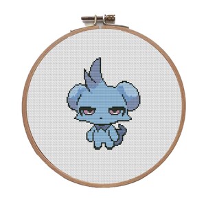 May include: A cross-stitch embroidery hoop featuring a light blue cartoon character with a grumpy expression. The character has large ears, a spiky head, and a small tail. The embroidery is set in a wooden hoop.