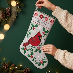 May include: A light gray Christmas stocking featuring a cross-stitched red cardinal bird perched on green holly branches with red berries. The stocking includes white snowflake accents and the name NOAH embroidered in black capital letters.