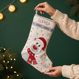 Op de afbeelding: Een crèmekleurige kerstsok met een in kruissteek geborduurde ijsbeer die een rode kerstmuts en een rode sjaal draagt. De sok is versierd met blauwe sneeuwvlokken en kleine rode hartjes, met de naam SOPHIA bovenaan geborduurd.
