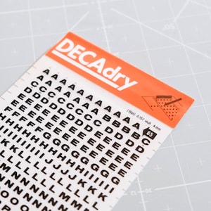 以下が含まれることがあります： 白い背景に黒い文字のステッカーが貼られたシート。文字はグリッド状に配置されており、AからOまであります。シートには「DECAdry」と表示され、文字のサイズがインチとミリメートルで記載されています。