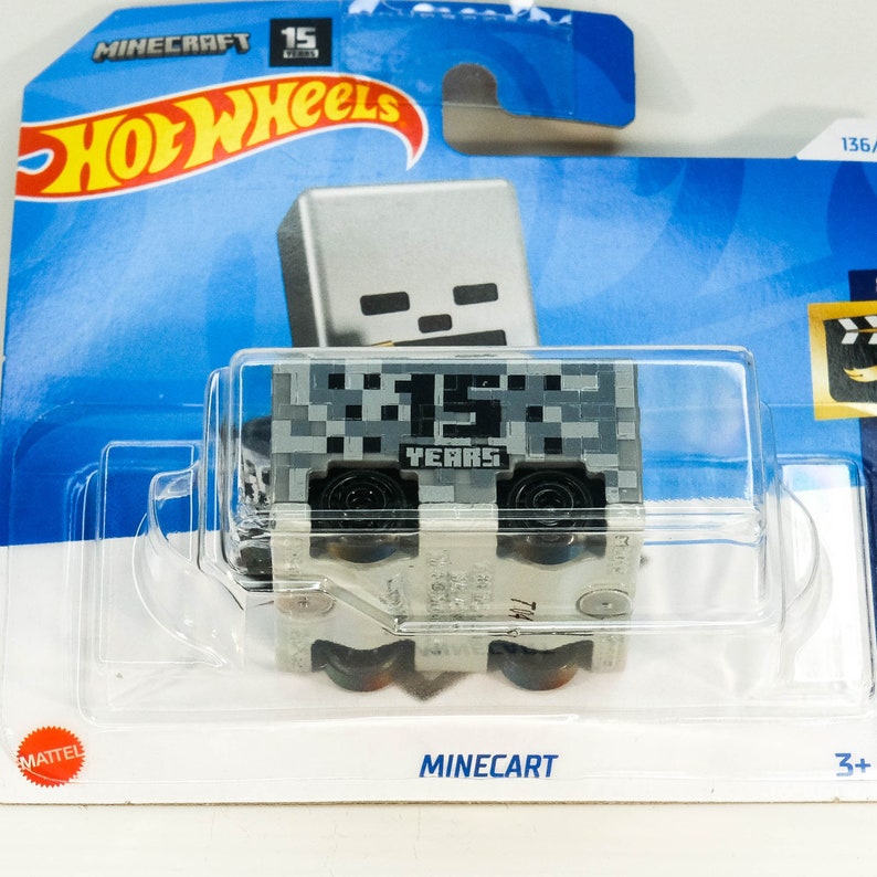 Puede incluir: Un coche de juguete Minecraft de color gris en forma de carro de minas con ruedas negras. El carro de minas tiene un dise&ntilde;o pixelado gris y negro con el texto "15 YEARS" en el lateral. El coche de juguete est&aacute; empaquetado en un fondo de cart&oacute;n azul y blanco con el logotipo de Hot Wheels y el logotipo de Minecraft.