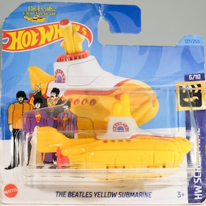 Könnte beinhalten: Ein Hot Wheels Spielzeug des Yellow Submarine der Beatles. Das gelbe U-Boot hat weiße und rote Akzente, mit einem Cartoon-Bild der Beatles auf der Seite. Die Verpackung enthält das Hot Wheels-Logo und den Text "THE BEATLES YELLOW SUBMARINE".