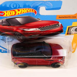 May include: Red toy car model of a Range Rover Velar with black trim and black wheels. The car is in a plastic package with the Hot Wheels logo and the text "hotwheels.com".
