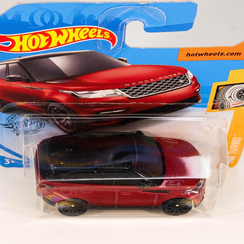 May include: Red toy car model of a Range Rover Velar with black trim and black wheels. The car is in a plastic package with the Hot Wheels logo and the text "hotwheels.com".