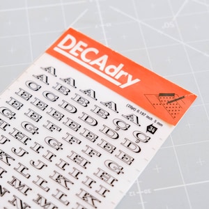 Può includere: Un foglio di adesivi con lettere Decadry con vari font. Gli adesivi sono in bianco e nero, con il nome del marchio in un banner rosso e bianco. Il foglio include le lettere dalla A alla N, con misure di 5 mm.