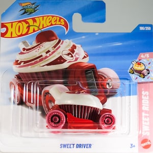 May include: A Hot Wheels Sweet Driver toy car in its original packaging. The car is designed to look like a cupcake with a red base, white frosting, and a cherry on top. The packaging features blue graphics, the Hot Wheels logo, and text.