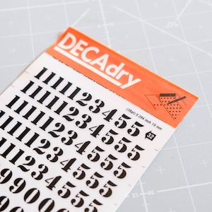 以下が含まれることがあります： 白い背景に黒い数字のステッカーが貼られたシート。数字は1から5まであり、高さは約10mm。シートには「DECAdry」と表示され、サイズ「32」と寸法「(38pt) 0.394 inch 10 mm」が記載されています。