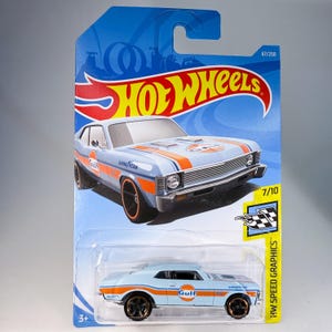 May include: A light blue Hot Wheels die-cast car with orange racing stripes and Gulf Oil branding. The car is a Chevrolet Nova, with black wheels and the text "GOOD YEAR" on the side. The packaging includes the Hot Wheels logo and "HW Speed Graphics".