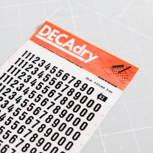以下が含まれることがあります： 白い背景に黒い数字のステッカーが貼られたシート。数字は1から9まであり、行に沿って繰り返されています。シートには「DECAdry」と「Made in Belgium」のラベルが貼られています。シートの幅は0.315インチ、厚さは8mmです。