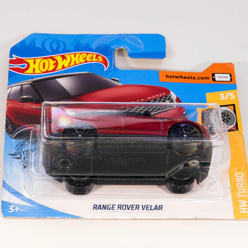 May include: Red Range Rover Velar toy car in blue and orange Hot Wheels packaging. The car is in a plastic case and the packaging has the text "Hot Wheels", "Range Rover Velar", "HW Turbo", and "hotwheels.com".