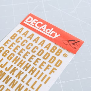 以下が含まれることがあります： 白いシートに赤と黒のDECAdryラベルが付いた、金色のアルファベット文字のステッカー。文字は大文字で、A、B、C、D、E、F、G、H、I、J、K、L、M、N、O、P、Q、R、S、T、U、V、W、X、Y、Zが含まれています。