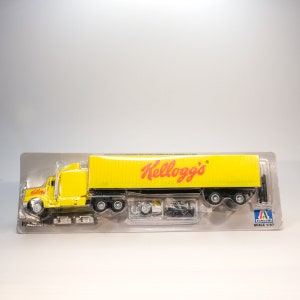 May include: A yellow toy semi-truck with a Kellogg's logo on the trailer. The truck is in a plastic package with a white background. The package says "Scale 1:87".