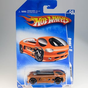 Op de afbeelding: Oranje en gouden Hot Wheels Deora II speelgoedauto op een blauwe en witte kaart. De auto heeft vlammen aan de zijkant en een zwart interieur. De kaart heeft het Hot Wheels logo en de tekst "Deora II" en "HW Designs".