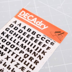May include: A sheet of black letter stickers on a white background. The letters are in a bold, sans-serif font. The sheet is labeled "DECAdry" and includes the size of the letters: (24pt) 0.276 inch 7 mm.