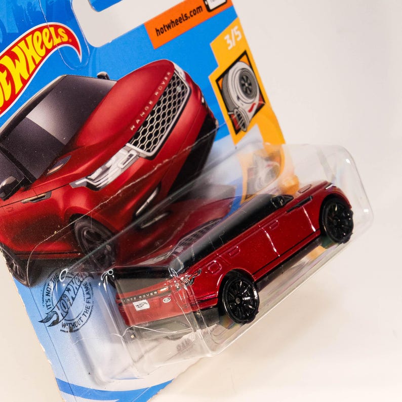 May include: Red toy car model of a Range Rover with black wheels and a black roof. The car is on a blue and orange background with the words "Hot Wheels" and "It's not just a toy, it's a flame without the flame".