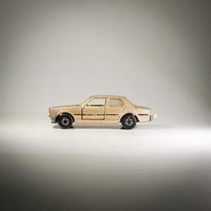 Könnte beinhalten: Ein beigefarbenes Spielzeugauto mit schwarzen Rädern und Zierleisten. Die Miniatur-Limousine hat ein klassisches Design mit einer viertürigen Karosserie und detaillierter Front und Heck. Das Auto steht vor einem weißen Hintergrund.
