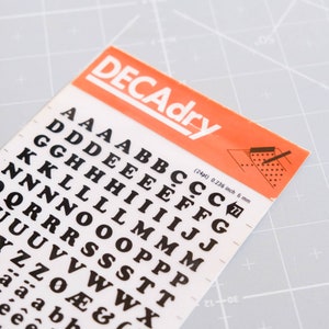 May include: A sheet of black letter stickers on a white background with the text "DECAdry (24pt) 0.236 inch 6 mm" in orange. The letters include A, B, C, D, E, F, G, H, I, J, K, L, M, N, O, P, Q, R, S, T, U, V, W, X, Y, Z, Æ, & and a.