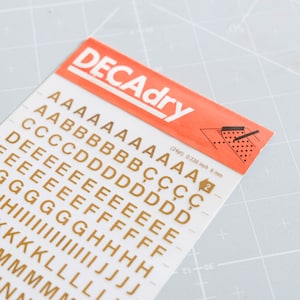 May include: A sheet of gold-colored Decadry dry transfer letters in various fonts and sizes. The sheet includes the text "DECAdry" in red and white. The letters are in a grid pattern, with measurements of 0.236 inch and 6 mm.