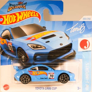 Może przedstawiać: Niebieski i żółty model samochodu Hot Wheels z odlewu ciśnieniowego, Toyota GR86 Cup, z napisami "Hot Wheels" i "Roman Empire 70" na boku. Samochód znajduje się na niebiesko-białej kartce z napisami "Toniz" i "HWJ-IMPORTS".