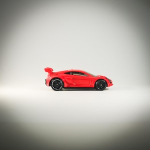 Puede incluir: Un coche de juguete rojo con ruedas negras y un alerón. El coche es un coche deportivo con un diseño elegante.