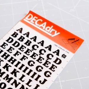以下が含まれることがあります： さまざまなフォントの黒いDecadryアルファベット文字のシート。文字は白い背景に印刷され、「DECAdry」と書かれたオレンジ色のヘッダーが付いています。シートにはさまざまなサイズの文字が含まれており、高さは9mmです。