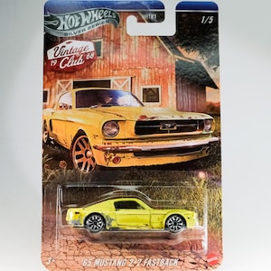 Puede incluir: Un coche de juguete fundido a presión amarillo '65 Mustang 2+2 Fastback de la serie Hot Wheels Silver. El embalaje presenta un diseño vintage con un granero de fondo y el texto "Vintage Club 68".