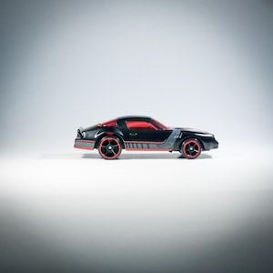 Könnte beinhalten: Ein schwarzes Spielzeugauto mit roten Fenstern und rot umrandeten Rädern. Das Fahrzeug hat graue Seitenstreifen und ein flaches Design. Die Karosserie ist schnittig mit einem Heckspoiler und einem kleinen Hot Wheels Logo an der Seite.