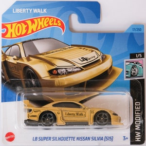 Puede incluir: Un coche de juguete de fundición a presión Hot Wheels de color dorado, un Nissan Silvia (S15) con las marcas "LB Super Silhouette" y "Liberty Walk". El coche tiene ruedas negras y un alerón negro. El coche está sobre un fondo azul y blanco con el logotipo de Hot Wheels.