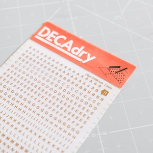 以下が含まれることがあります： オレンジ色のラベルに白い文字で「DECAdry」と書かれた、金色のアルファベット文字ステッカーのシート。クラフトやスクラップブッキングのプロジェクトに最適な、さまざまなサイズの文字が含まれています。
