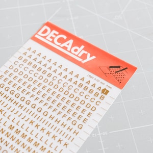 以下が含まれることがあります： さまざまなサイズの金色のDecadryドライ転写文字のシート。文字は白い背景に、行と列に配置されています。シートの上部には、「DECAdry」のブランド名が記載された赤いバナーがあります。