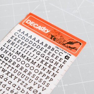 Peut inclure: Une feuille d'autocollants de lettres noires sur fond orange. Les lettres sont de différentes tailles et polices, y compris les lettres majuscules et minuscules, les chiffres et les symboles. La feuille est étiquetée "DECAdry" et comprend le texte "(20pt) 0.197 inch 5 mm 85".