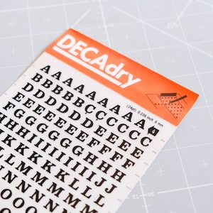 May include: A sheet of black letter stickers on a white background. The letters are in a variety of sizes, from A to O. The sheet is labeled "DECAdry" and includes the text "(24pt) 0.236 inch 6 mm".