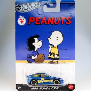 以下が含まれることがあります： 青い黄色のホットウィールダイキャストカー。側面にピーナッツのキャラクターが描かれています。この車は1985年のホンダCR-Xです。パッケージには、ピーナッツコミックのチャーリーブラウンとルーシーの漫画が描かれています。